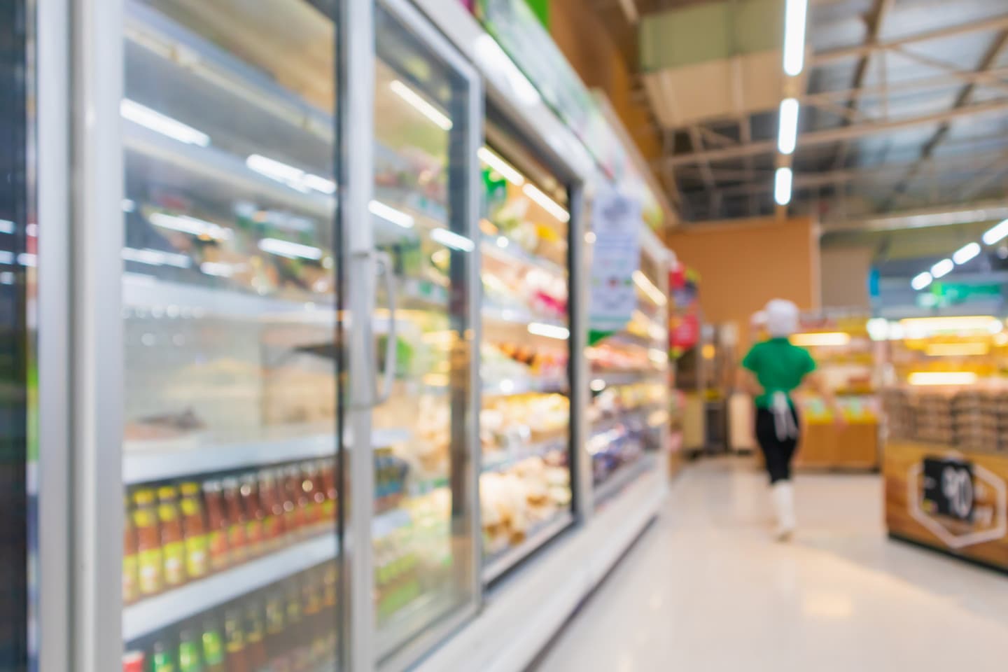 Blurred image of a supermarket aisle showcases refrigerated shelves on the left, stocked with various products. A person in green uniform, perhaps from commercial refrigeration repair, is seen walking in the background.