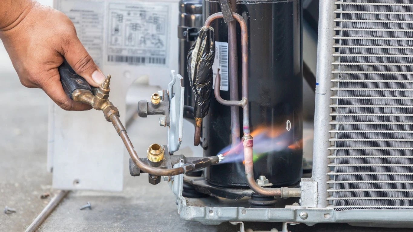 A person expertly uses a welding torch on metal pipes connected to an air conditioning unit, similar to a skilled technician tackling furnace repair in Mississauga, Furnace Issues