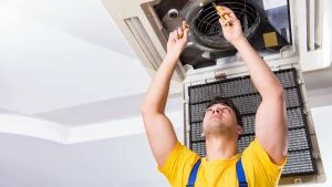 A technician in a yellow shirt and blue overalls is meticulously conducting AC maintenance, skillfully repairing a ceiling air conditioning unit with precision tools, Energy-Efficient HVAC Systems