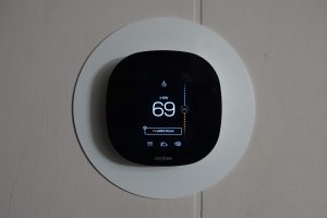 A digital thermostat with a black interface, expertly installed, displays the temperature set to 69 degrees. It is mounted on a circular white wall plate, Thermostat Installation