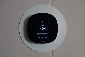 A digital thermostat with a black interface, expertly installed, displays the temperature set to 69 degrees. It is mounted on a circular white wall plate, Thermostat Installation