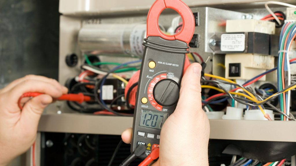 Close-up of a person using a digital clamp meter to measure voltage in a circuit with wires and electrical components, much like the detailed care needed for furnace repair in Mississauga and Brampton.