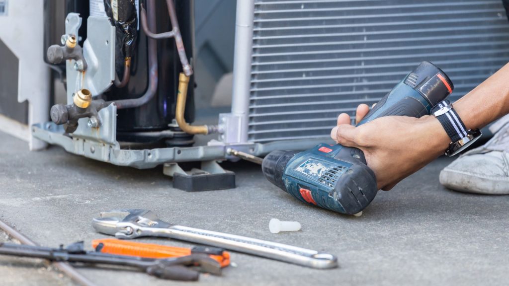A person is skillfully using a power drill to repair an air conditioning unit, expertly managing various tools scattered on the ground nearby, showcasing their proficiency in HVAC systems.