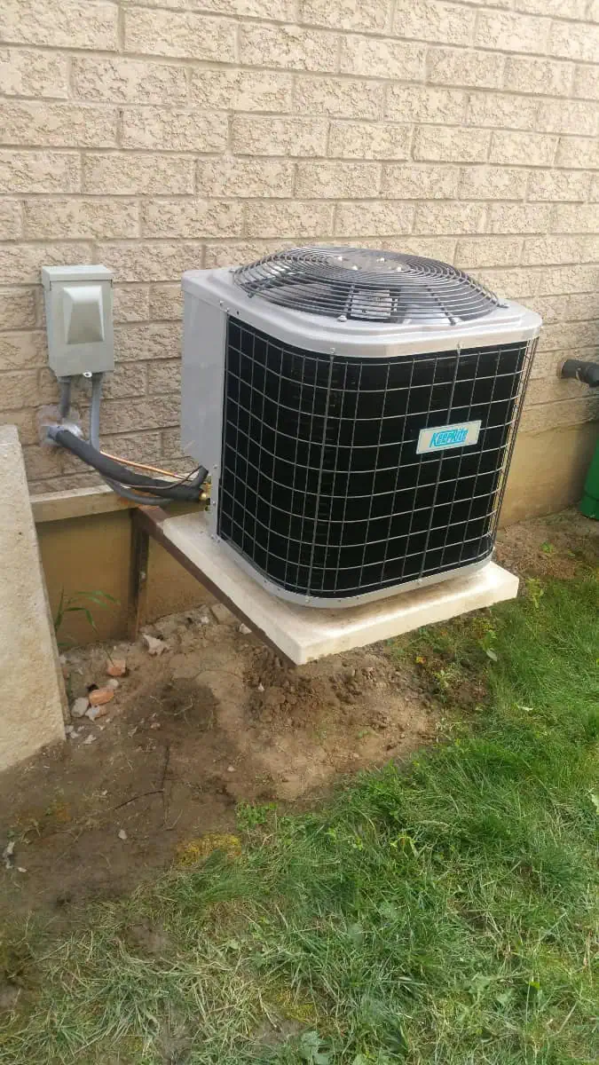 Air conditioning repair Brampton specialists ensure that the unit, installed on a concrete platform outside a brick building with connected electrical components, runs efficiently. Lush grass and rich soil lie in the foreground, enhancing the serene setting.