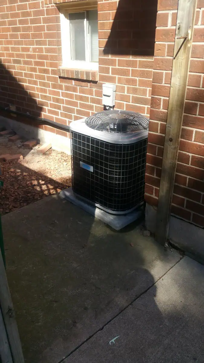 Outdoor air conditioning unit against a brick wall, near a window, on a concrete slab. If you need air conditioning repair in Brampton for this setup, reliable services are available to ensure it functions efficiently.