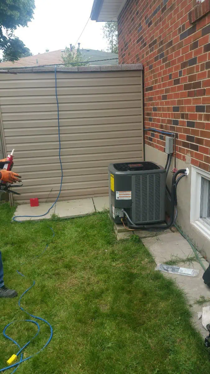 An air conditioning unit sits securely outside a brick house on a concrete slab, with a shed in the background. On the left, a person holds a blue hose, illustrating the process of air conditioning repair in Brampton.