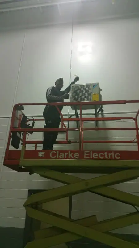 A person stands on a lift, skillfully performing maintenance on a wall-mounted Clarke Electric unit indoors. This routine task ensures that the HVAC system in Oakville operates smoothly and efficiently, keeping the environment comfortable year-round.