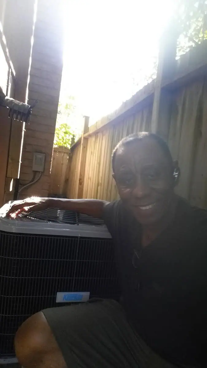 A man smiles beside an air conditioning unit in a narrow outdoor space, sunlight filtering through the fence, showcasing his expertise in air conditioning repair Brampton.