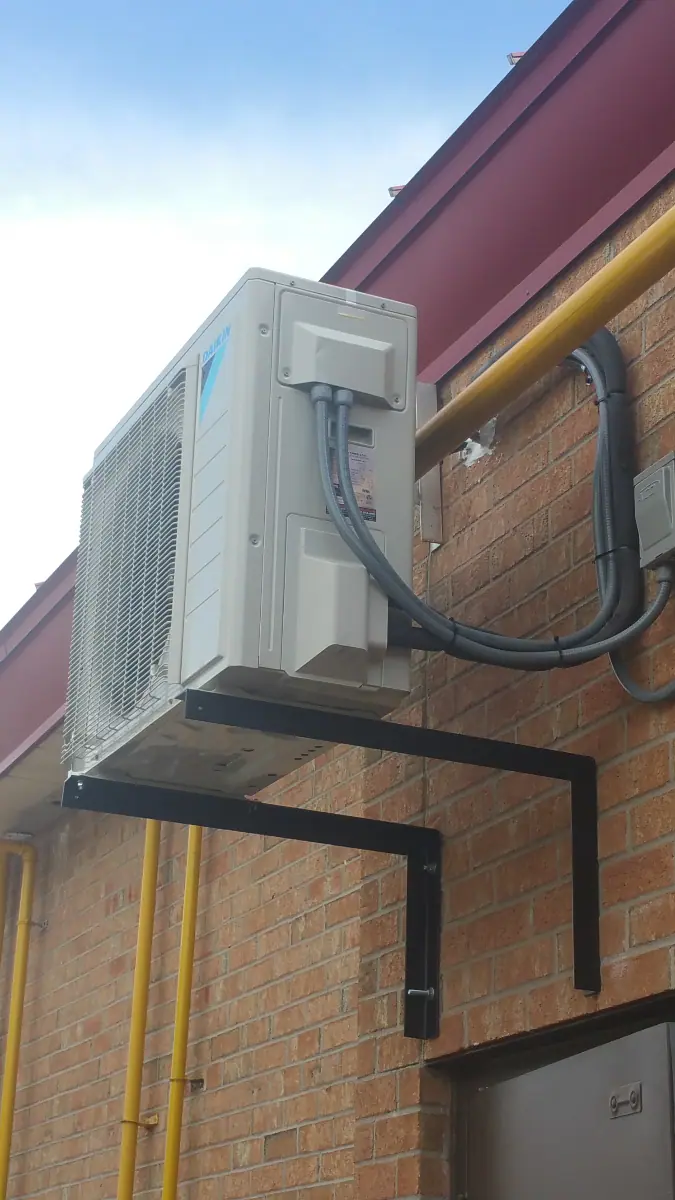 A wall-mounted outdoor air conditioning unit with cables runs along the brick exterior of a building, showcasing the skilled craftsmanship typical of air conditioning repair in Brampton.