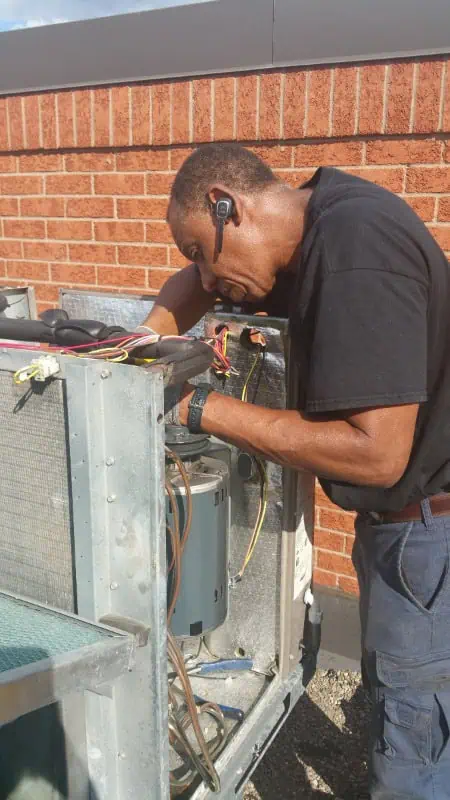 A skilled technician from HVAC Oakville is on a rooftop, wearing a headset as he meticulously repairs an outdoor air conditioning unit, framed by brick walls in the background.