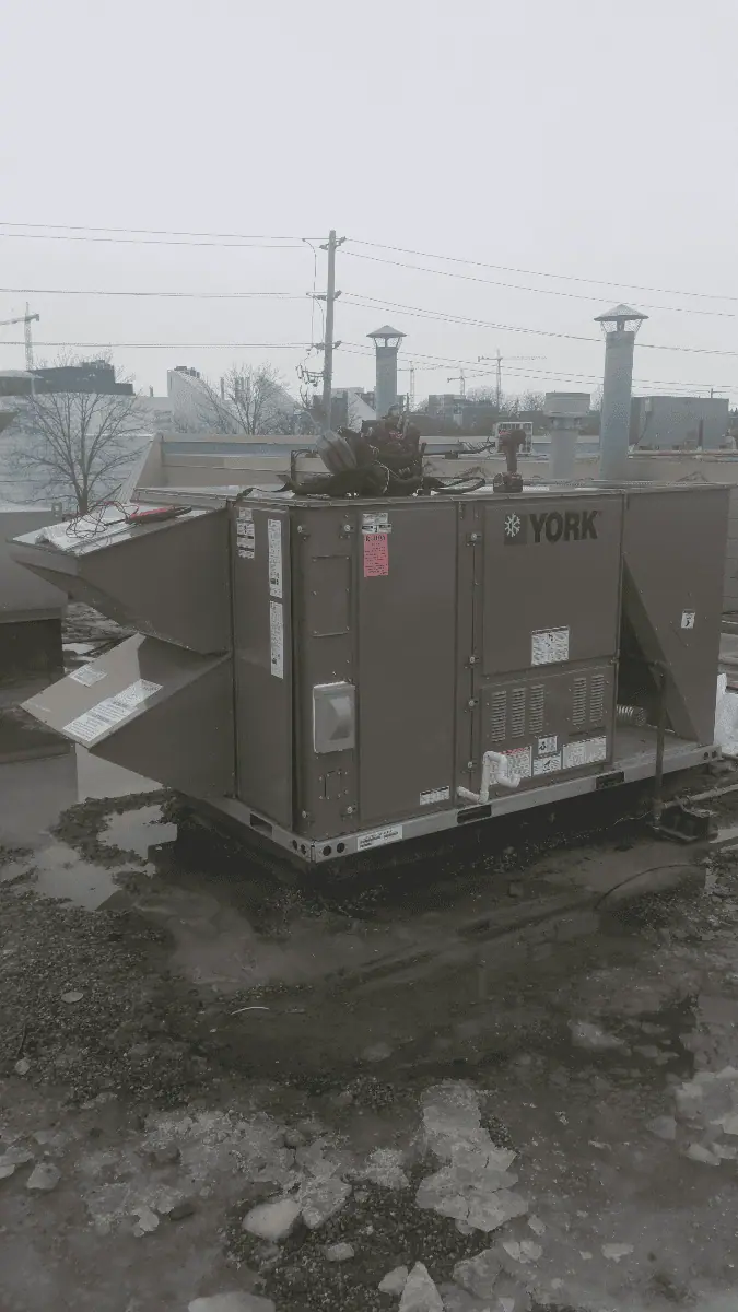 A large industrial HVAC unit with visible ducts and panels sits outdoors on a gravel surface, reminiscent of Mississauga furnace repair setups. Several buildings and power lines are visible in the background, conveying an urban industrial scene.