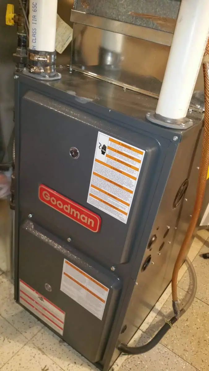 A gray Goodman furnace, installed in a utility area, boasts attached pipes and visible safety stickers. For those in need of Mississauga furnace repair, this reliable model ensures efficient operation all winter long.