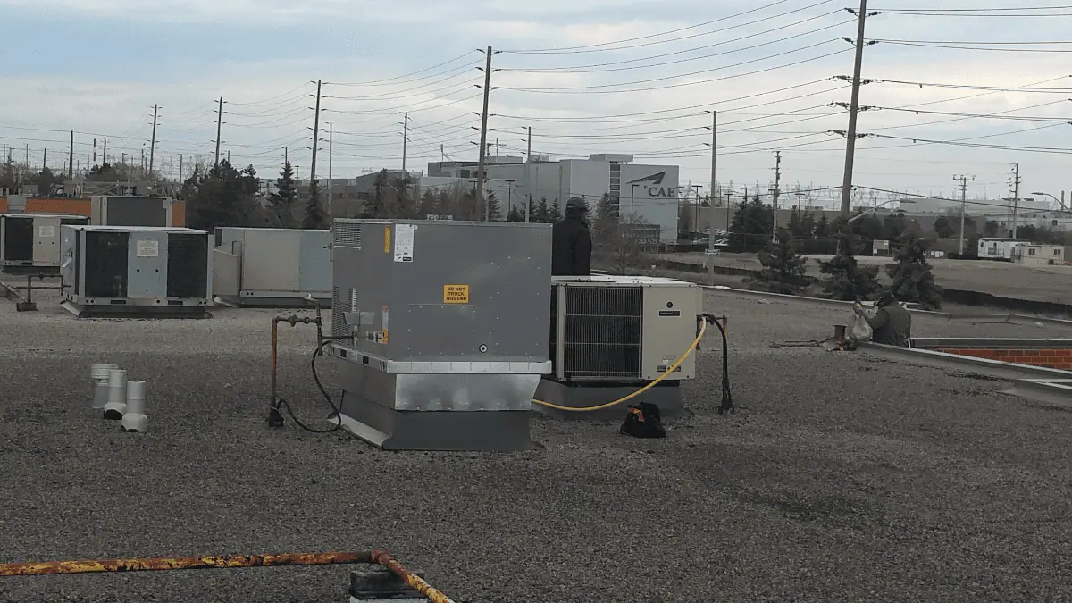 A person stands on a flat rooftop near HVAC units, representing an experienced HVAC contractor in Brampton, Canada. They're surrounded by industrial buildings and a cloudy sky in the background.