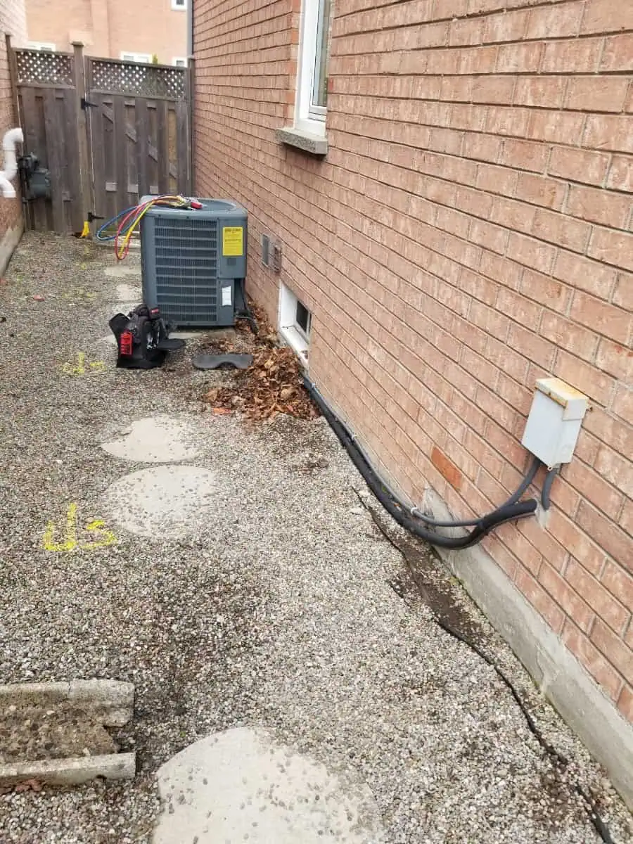 A narrow alley with a brick wall and an HVAC Milton unit, scattered leaves, and a gravel path dotted with circular stepping stones.