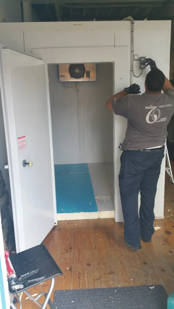 A skilled technician is providing HVAC service in Halton Hills, meticulously working on the wiring of an industrial refrigeration unit. The door is open, revealing a blue and gray interior with a cooling unit above.