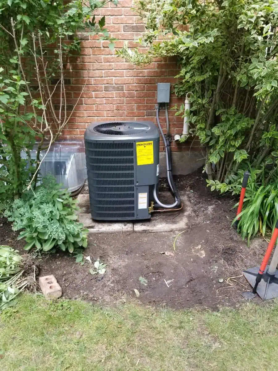 Air conditioning unit installed on a concrete pad next to a brick wall, surrounded by plants and tools on the grass, showcasing the quality work of HVAC service Halton Hills.