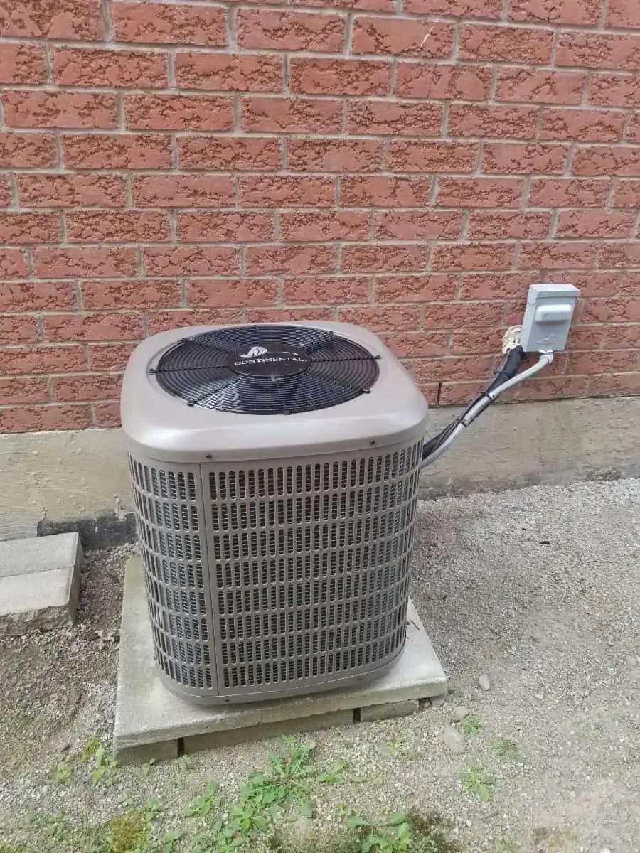 An outdoor HVAC unit in Milton rests on a concrete slab beside a brick wall, with its electrical connection clearly visible.