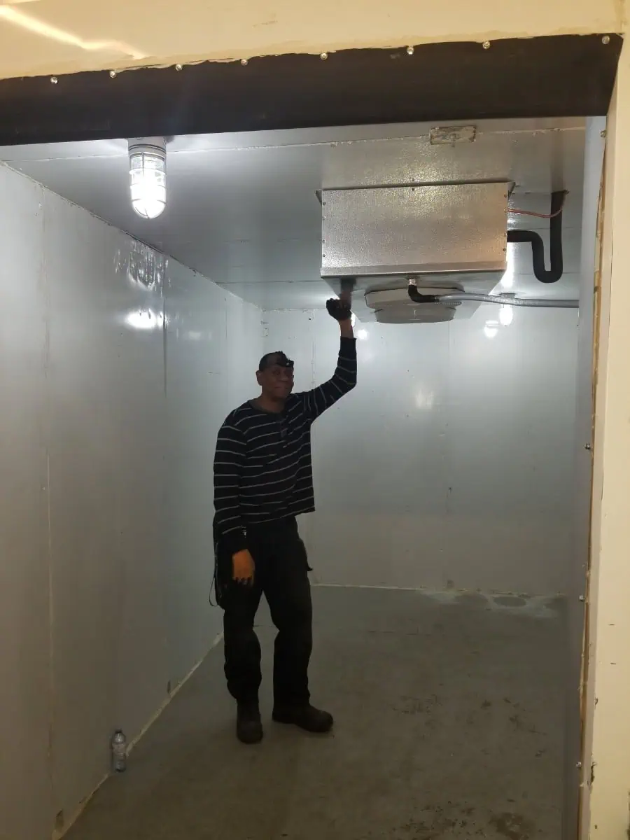 A person stands in a spacious, empty, white-walled room, wearing a striped shirt and black pants. With one hand gently touching an HVAC Milton ceiling vent, they appear contemplative amidst the minimalist setting.