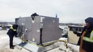 Two workers are inspecting a large rooftop HVAC unit on a snowy day in Brampton. One, wrapped up in a hooded jacket, holds a tablet to ensure the Emergency Heating Repair is on track.